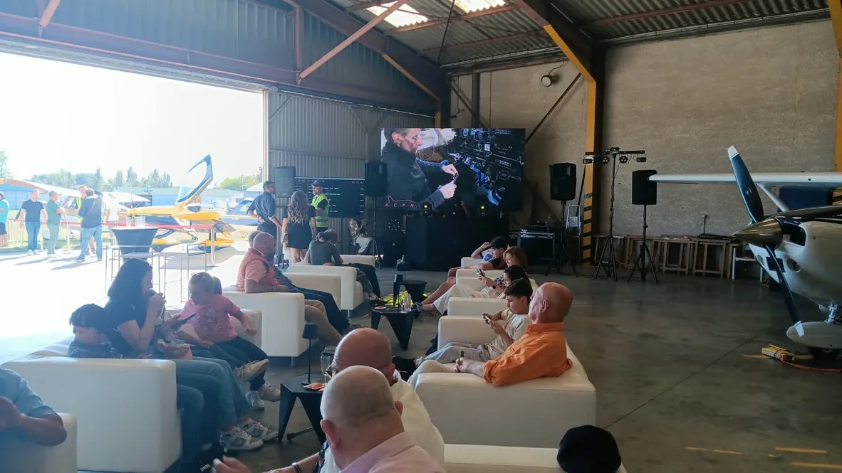 Visiteurs installés dans les canapés du hangar devant un écran géant diffusant une vidéo de vol, lors du Fly-in